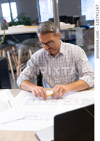 Male architect reviewing blueprints at modern open-plan office desk, with laptop and pencil 128741167