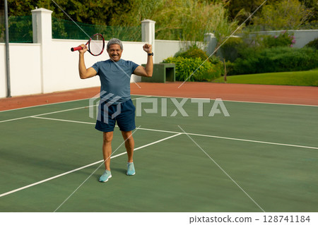 Senior man raising arms celebrating on outdoor tennis court, holding red-and-white racket Senior man raising arms celebrating on outdoor tennis court, holding red-and-white racket 128741184