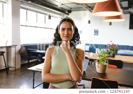 Woman standing thoughtfully in modern coworking office, surrounded by laptop, notebooks and plants 128741197