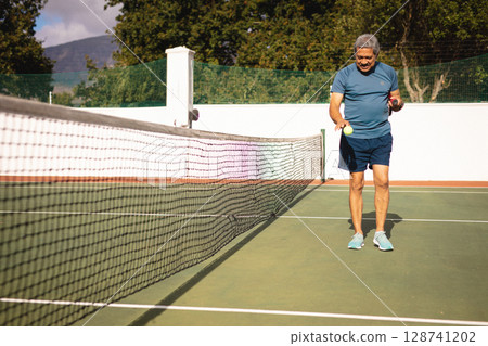 Senior man picking up yellow tennis balls near net on outdoor hard court, copy space 128741202