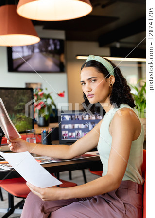 Woman reviewing printed sheets at open?plan office desk, surrounded by monitors and red chairs Woman reviewing printed sheets at open?plan office desk, surrounded by monitors and red chairs 128741205