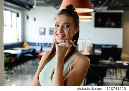 Woman standing smiling in modern open?plan office, with pendant lamps and lounge bench 128741210