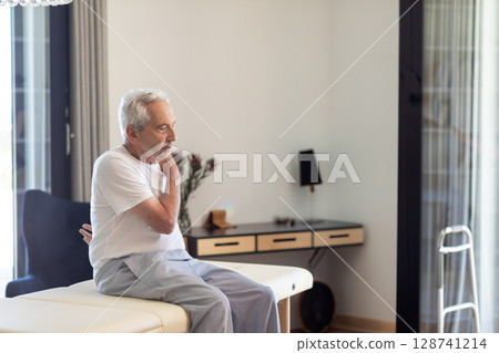 Senior man sitting on therapy table in therapy room holding painful neck with walker, copy space 128741214