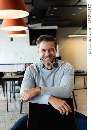 Male office worker sitting backwards on chair in open office under orange lamps, smiling at camera Male office worker sitting backwards on chair in open office under orange lamps, smiling at camera 128741217