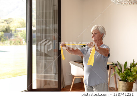 Senior woman performing resistance band exercise at home near glass doors, holding yellow band 128741221