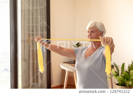 Senior woman stretching yellow resistance band in living room by sliding doors, with potted plants 128741222