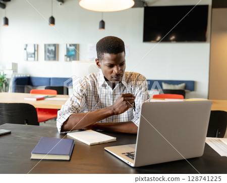 African American man using silver laptop at coworking table with notebooks, pen and smartphone 128741225