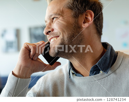 Mid adult man talking on smartphone inside modern interior, wearing knit sweater and smiling Mid adult man talking on smartphone inside modern interior, wearing knit sweater and smiling 128741234