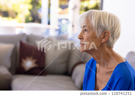 Senior woman sitting on gray sofa in living room, with large windows, burgundy pillow, copy space 128741251