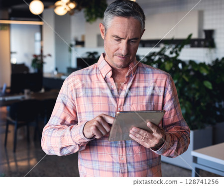 Middle-aged man using tablet computer in modern open-plan office space, with desks and plants 128741256