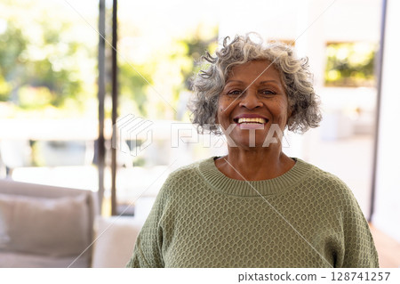 Senior woman standing by sofa in living room, admiring daylight from windows and plants, copy space 128741257
