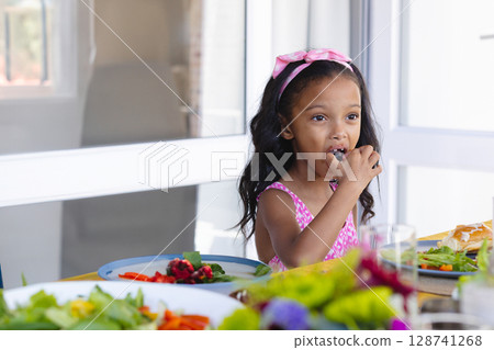 Girl eating berry at dining table in bright modern room, with colorful fruit plates, copy space 128741268