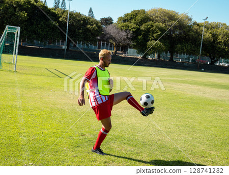 Male soccer player controlling soccer ball on grass soccer field, with goalpost and fluorescent bib Male soccer player controlling soccer ball on grass soccer field, with goalpost and fluorescent bib 128741282