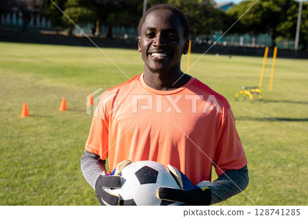 Teenage boy standing smiling on soccer field, near cones, wearing gloves and holding soccer ball 128741285