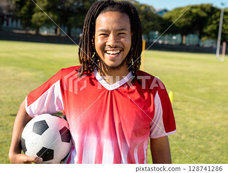 Young adult man standing on grassy sports field during soccer practice, holding soccer ball 128741286