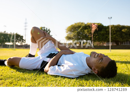 Male soccer player lying on soccer pitch grass near goal frame clutching knee in white jersey Male soccer player lying on soccer pitch grass near goal frame clutching knee in white jersey 128741289