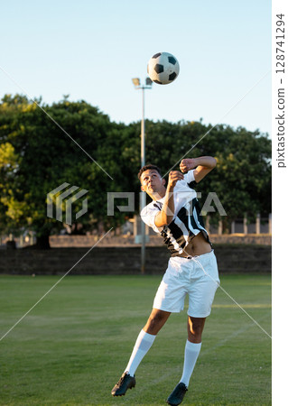 Young adult male athlete leaping in mid-air on grass soccer pitch, heading soccer ball Young adult male athlete leaping in mid-air on grass soccer pitch, heading soccer ball 128741294