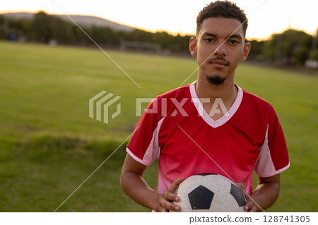 Young adult male holding soccer ball standing on soccer pitch near goalpost at dusk, copy space 128741305