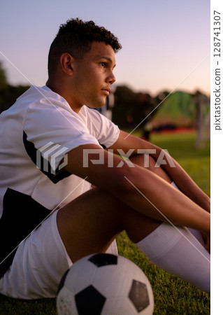 Male soccer player sitting on grassy pitch at sunset with soccer ball and goalposts, copy space 128741307