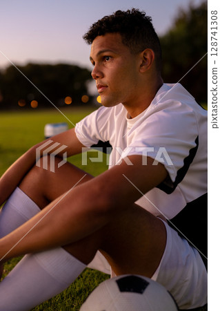 Young male soccer player sitting on grass field at dusk, gazing into distance with soccer ball 128741308