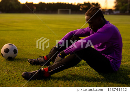 Teenage boy tightening goalkeeper gloves on grass soccer pitch at sunset, with ball cleats goalpost 128741312