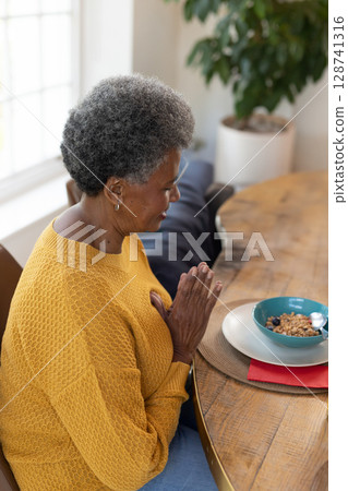 Senior African American woman praying at home dining table, with blueberry cereal, spoon, plant 128741316