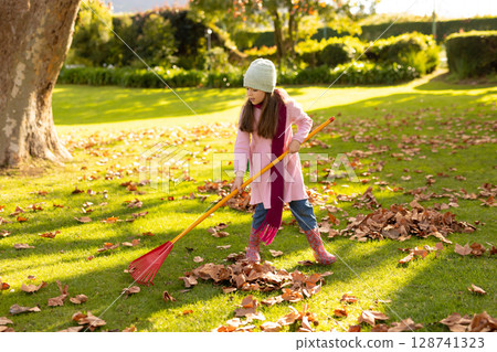 Girl child raking autumn leaves on sunlit garden lawn using orange-handled rake, copy space Girl child raking autumn leaves on sunlit garden lawn using orange-handled rake, copy space 128741323