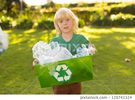 Boy holding green recycling bin in sunlit backyard, sorting plastic bottles and clear bags 128741324