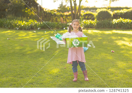 Girl holding green recycling crate filled with plastic bottles and paper scraps on sunlit backyard 128741325
