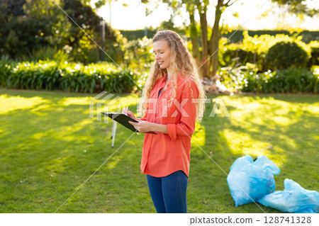 Woman standing and writing on black clipboard in sunny backyard with blue garbage bags, copy space 128741328