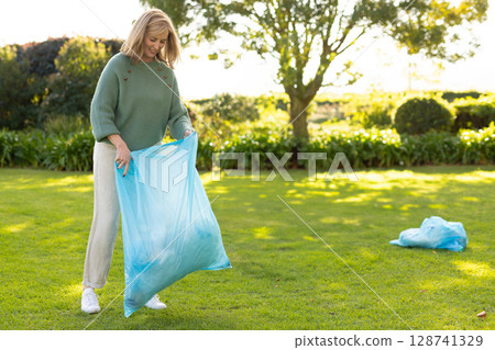 Woman holding open blue trash bag on suburban garden lawn, collecting yard debris, copy space Woman holding open blue trash bag on suburban garden lawn, collecting yard debris, copy space 128741329