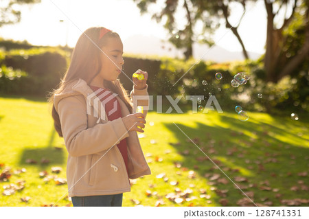 6-year-old girl blowing soap bubbles in garden, with yellow wand and solution bottle, copy space 128741331