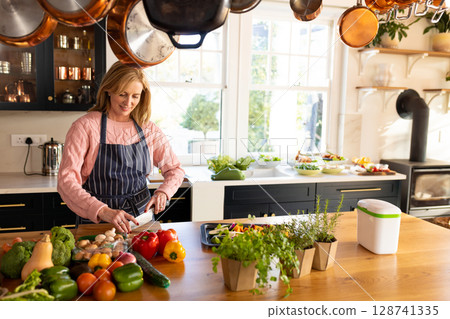 Mature woman slicing bell pepper on wooden cutting board in modern home kitchen with potted herbs Mature woman slicing bell pepper on wooden cutting board in modern home kitchen with potted herbs 128741335