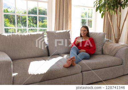 Young adult woman sipping coffee from white mug on gray sofa in living room, copy space 128741346