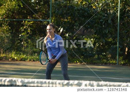 Young adult woman gripping tennis racket in ready stance on tennis court with net and fence 128741348