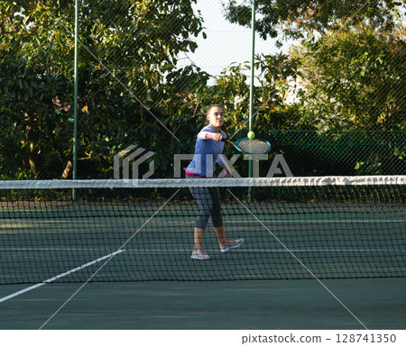 Young adult woman swinging racket and volleying tennis ball over net on outdoor court at park 128741350