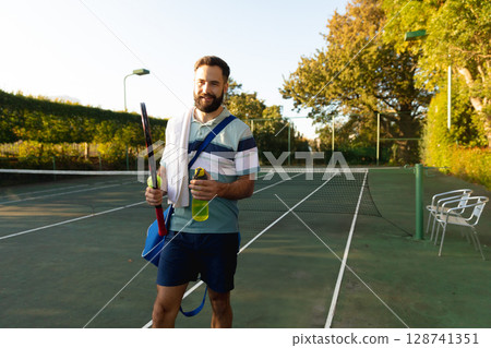 Man walking on outdoor tennis court, carrying racket, holding green water bottle and sports bag 128741351
