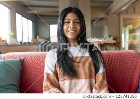 Teenage girl sitting and smiling on red sofa in living room, with books and plants 128741356