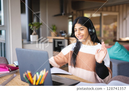 Teenage Middle Eastern girl sitting at table in open-plan home, waving at laptop with headphones 128741357