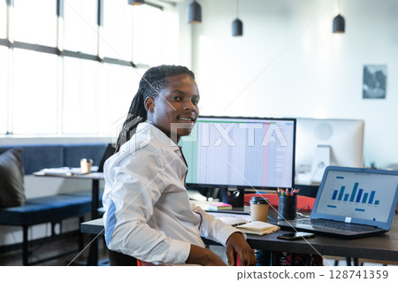 African American man working at desk in modern open-plan office, with laptop, monitor, coffee cup 128741359
