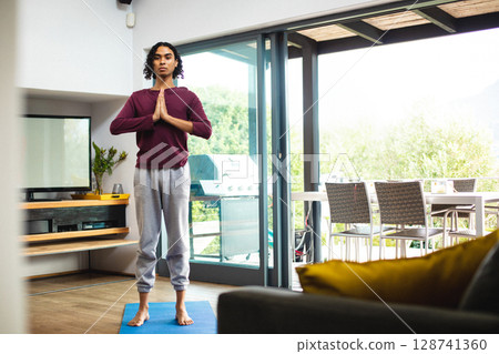 Non-binary person doing yoga prayer pose on blue mat in modern living room, by sliding doors 128741360