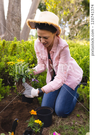 Mature woman kneeling in residential garden bed, planting flowering plant with trowel and pot 128741374
