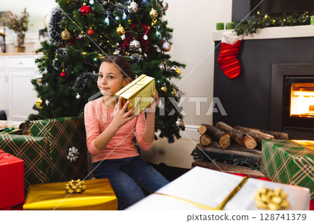 Female child sitting beside Christmas tree at home holding gold-wrapped present to ear, copy space 128741379