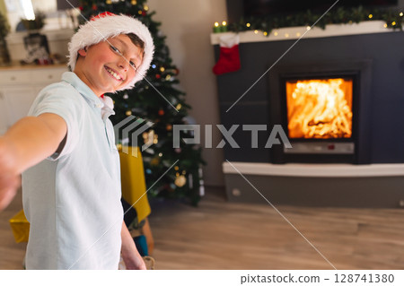 Boy standing at home with Christmas tree, Santa hat, stocking, garland lights, copy space Boy standing at home with Christmas tree, Santa hat, stocking, garland lights, copy space 128741380
