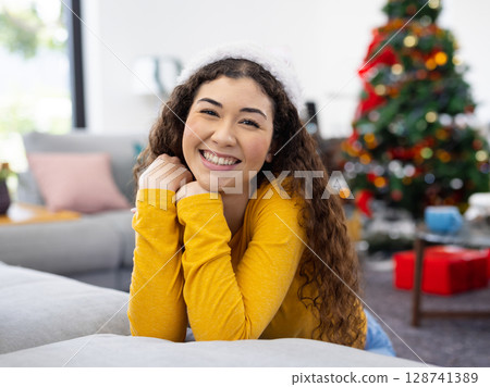 Young adult woman leaning on sofa in living room with Christmas decor, tree, pillows and headband 128741389