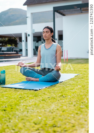 Woman sitting cross?legged in meditative pose on backyard lawn, with yoga mat and water bottle Woman sitting cross?legged in meditative pose on backyard lawn, with yoga mat and water bottle 128741390
