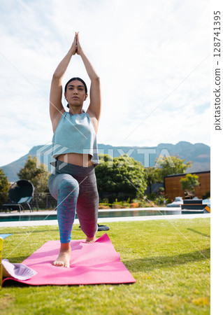Diverse transgender person practicing yoga lunge by pool on pink mat, wearing colorful leggings 128741395