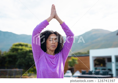 Woman doing yoga prayer pose outdoors on modern patio, with green shrubs and hills 128741397