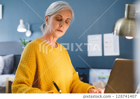 Senior woman writing on papers at bedroom office corner, with laptop, pen and desk lamp 128741407