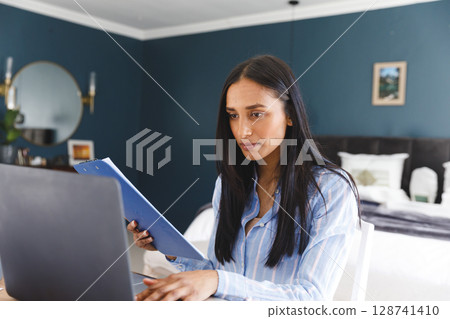 Diverse woman working at white desk in bedroom home office, using laptop and clipboard, copy space 128741410
